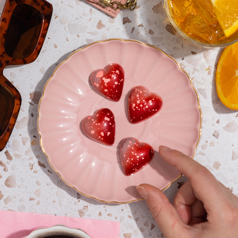 Four Cafe Amore chocolates on a pink plate. The chocolates are pink and heart-shaped. A hand reaches in as if to grab one off of the plate.  The plate sits on a white table surrounded by sunglasses and a drink with an orange slice.